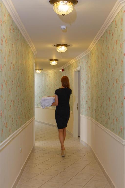 Sieviete, tērpta melnā kleitā, nesis kaudzi dvieļu pa gaišu, dekorētu viesnīcas koridoru. Koridors ir apgaismots ar elegantiem griestu lampām un greznu tapeti.