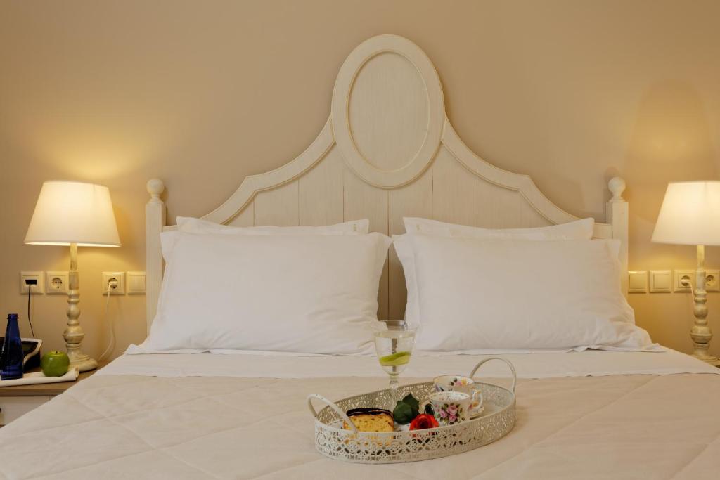 Mūsdienīgs un elegants guļamistabas interjers ar baltu gultas galvu un spilveniem, papildināts ar apgaismojumu un ērtu galdiņu, uz kura atrodas dzērienu un uzkodu pasniegšanas trauks.