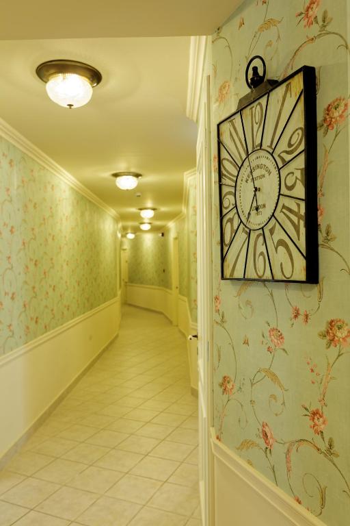 Elegants gaiteņš ar ziedu rakstu tapetēm un melnu pulksteni pie sienas, radot mājīgu atmosfēru. Gaismas avoti pie griestiem piešķir siltumu un komfortu.