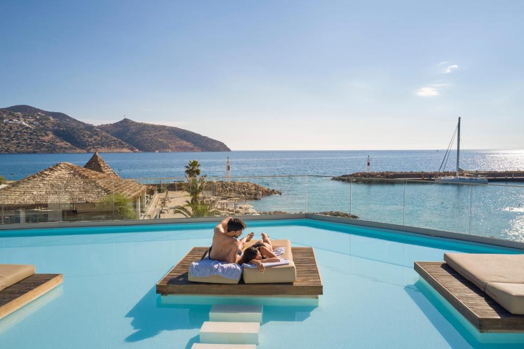 Pāris atpūšas pie baseina Mirabello Bay Luxury Resort, ar skatu uz piekrasti.