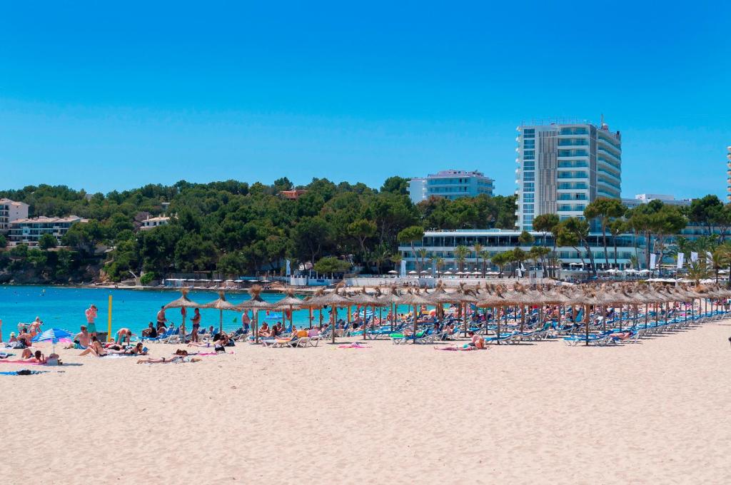 Skaists pludmales skats ar saulainu debesi, zilu ūdeni un cilvēkiem, kas atpūšas uz saules. Pludmalē ir daudz saulessargu un sauļošanās krēslu, kā arī apkārt esošās viesnīcas un zaļumi.
