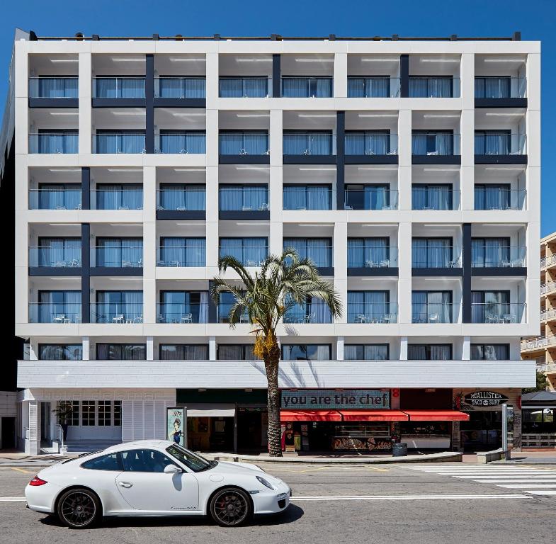 Mūsdienīga viesnīca ar baltu fasādi un lieliem logiem, priekšplānā redzama balta sporta automašīna un palmu koks.