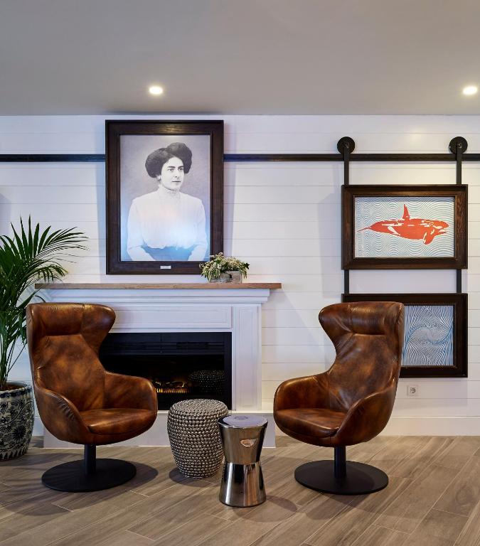 Mūsdienīgs interjers ar divām brūnām krēsliem, kam blakus ir kamīns un mākslas darbi uz sienas, tostarp melnbalta portrets un krāsains zīmējums.