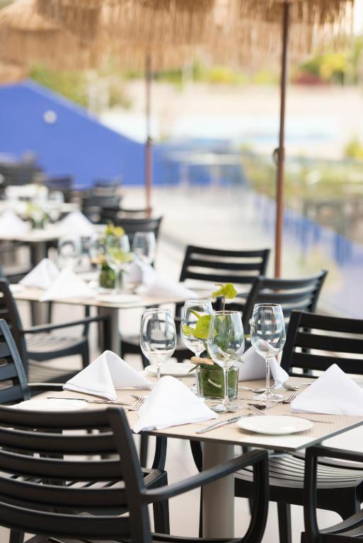 Skaisti galdiņi ar glāzēm un puķēm, sagatavoti ēdināšanas vietai, ar saulessargiem un baseina fonā.