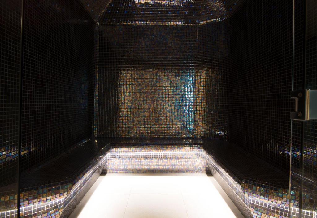 Mūsdienīgs tvaika pirts interjers ar spoguļojošām mozaīkas flīzēm, kas rada elegantu un relaksējošu atmosfēru. Apgaismojums akcentē telpas dizainu.