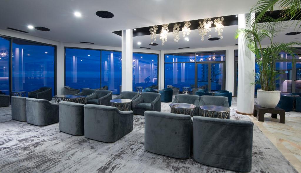 Mūsdienīgs atpūtas telpas interjers ar pelēkām mēbelēm, stikla sienām un skatu uz jūru. Telpā ir eleganti galdi un augi, radot relaksējošu atmosfēru.