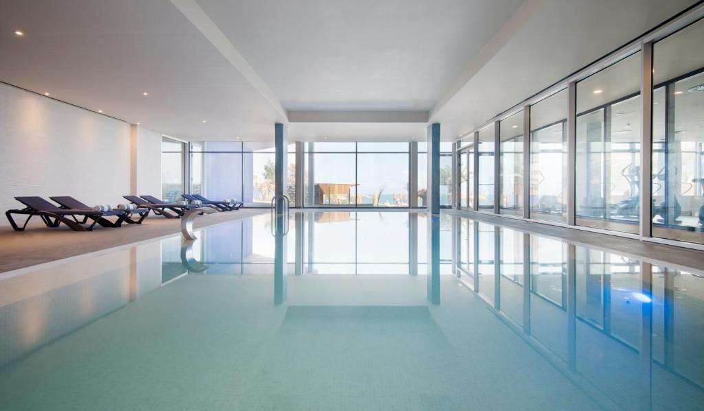 Iekštelpu peldbaseins ar modernu dizainu, kurā redzami atpūtas krēsli un lieli logi, kas nodrošina dabiska apgaismojuma plūsmu. 