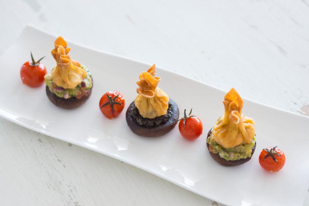 Trīs pildītas sēnes uz baltas šķīvja, dekorētas ar kraukšķīgām uzkodām un apvienotas ar grauzdētiem tomātiem. Svaiga un estētiska uzkoda, ideāli piemērota svinībām.