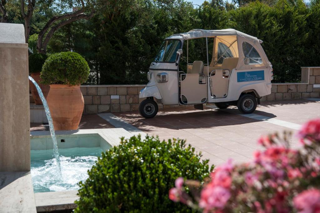 Balta tuk-tuk automašīna stāv pie ūdens strūklakas, fonā redzami zaļi krūmi un puķu dobes ar ziediem.