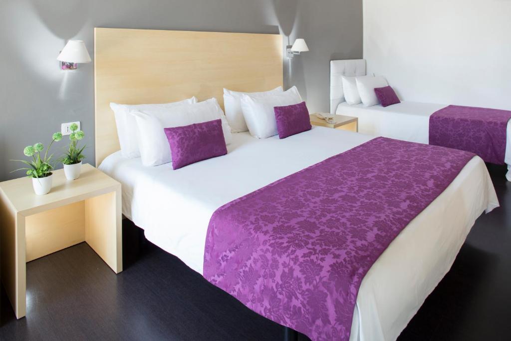 Moderns viesnīcas numurs ar divām gultām, rotāts ar purpura krāsas gultas pārklājiem un spilveniem, blakus stūra galdiņš ar augiem.