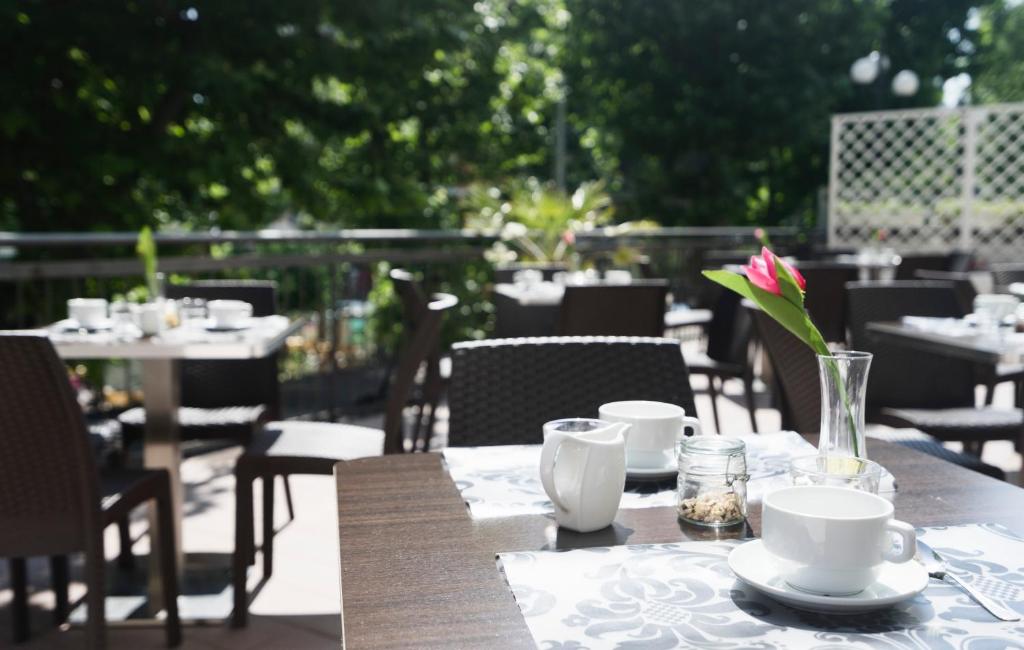 Terases restorāna skats ar galdiem, kafijas tases un ziedu dekorāciju. Daba un zaļumi veido patīkamu atmosfēru.