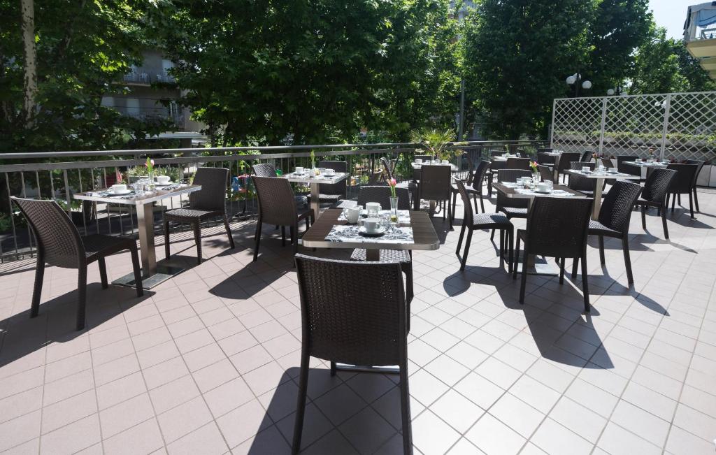 Āra terase ar galdiem un krēsliem, kas sakārtoti piekļūšanai saules gaismai un dārza ainavai. Galdiem ir novietotas salvetes un trauki, radot viesmīlīgu atmosfēru.