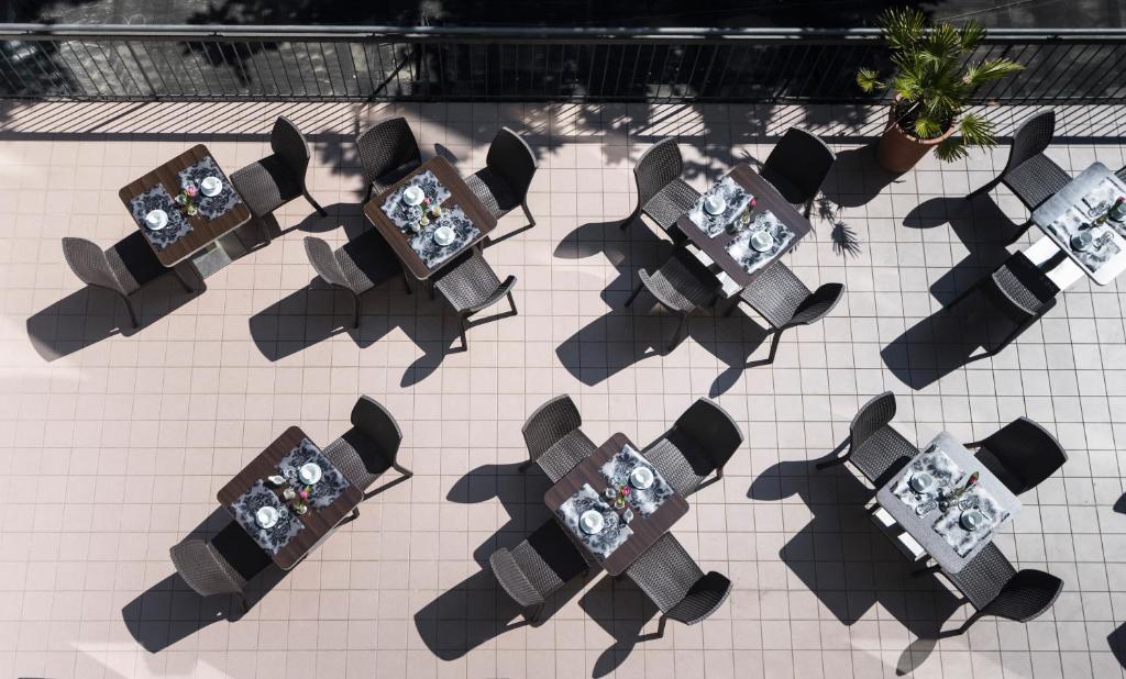 Āra kafejnīcas galdi ar krēsliem, sakārtoti uz flīžu grīdas, ar galdu uzklājumiem un ziediem. Gaismas un ēnu spēles rada patīkamu atmosfēru.