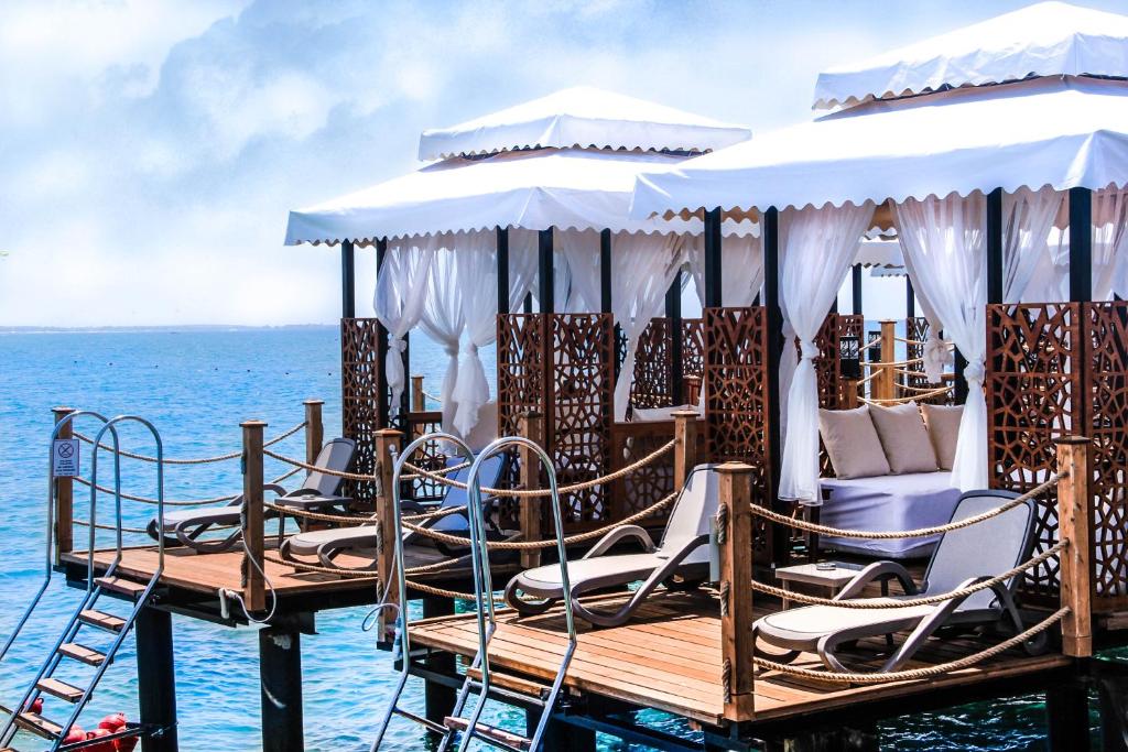 Koka platforma ar ērtām guļvietām un baltiem nojumiem, kas atrodas virs ūdens, piedāvā relaksējošu atmosfēru. Fons ir skaidrs, zils ūdens un debesu ainava.