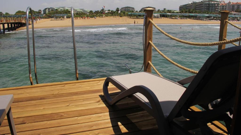 Koka terase ar saulessargu un skatu uz pludmali, kur viļņi maigi krāso smiltis. Fons ietver viesnīcas un viesu namus, radot relaksējošu pludmales atmosfēru.