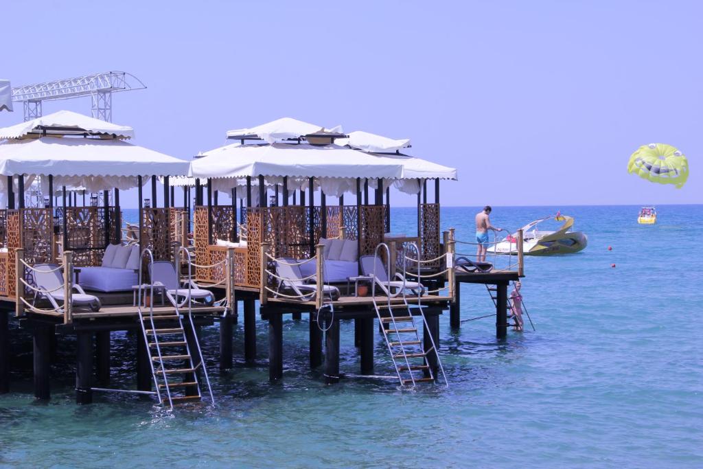 Tropu pludmales kafejnīca ar koka platformām virs ūdens, kur cilvēki atpūšas un piedalās ūdens aktivitātēs, piemēram, parasailing. Skaists zils ūdens un saulains laiks.