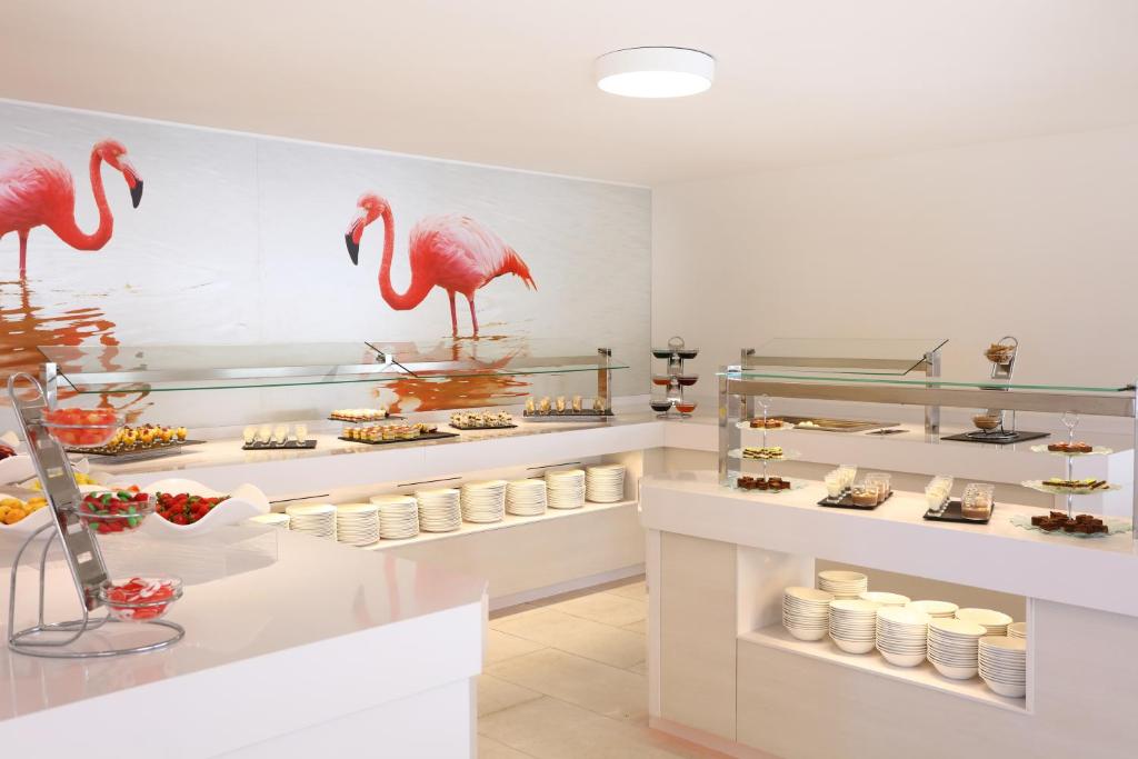 Kafeterijas stūrītis ar elegantu baltu dizainu un daudzveidīgu uzkodu izvēli, fona attēlā redzami flamingi.