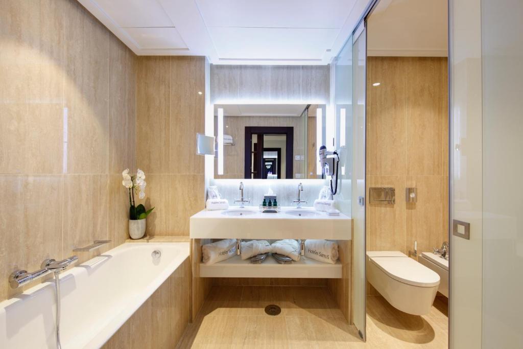 Mūsdienīgs vannas istabas interjers ar elegantu dizainu, ietverot vannu, divus izlietnes un lielu spoguli. Kvalitatīvi materiāli un apgaismojums rada patīkamu atmosfēru.