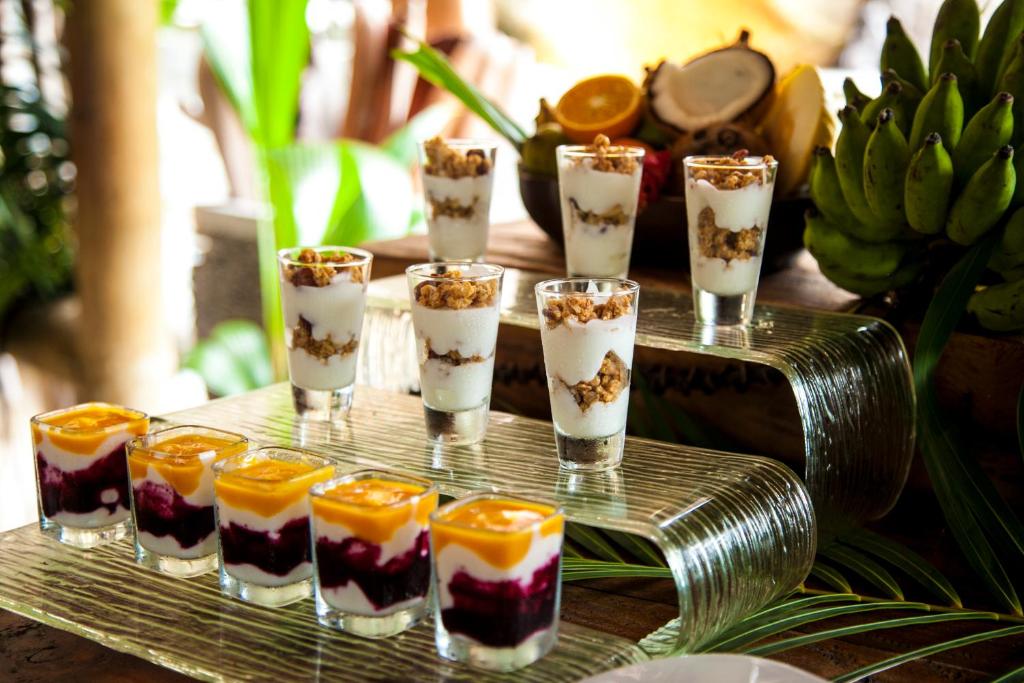 Deserta trauku kolekcija ar jogurtu, granolu un augļu mērci, kas pasniegti stilīgā stikla plauktā. Fons papildināts ar tropiskiem augļiem un zaļumiem.