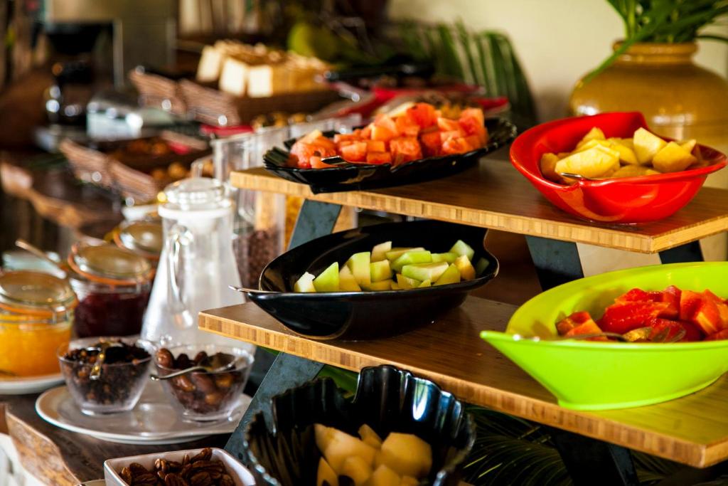 Svaigu augļu bufete ar dažādām krāsainām bļodām, kas satur melones, ananasus un citus augļus, izvietota uz koka plauktiem. Fonā redzami burciņas ar ievārījumiem un kafijas automāts, radot patīkamu brokastu atmosfēru.