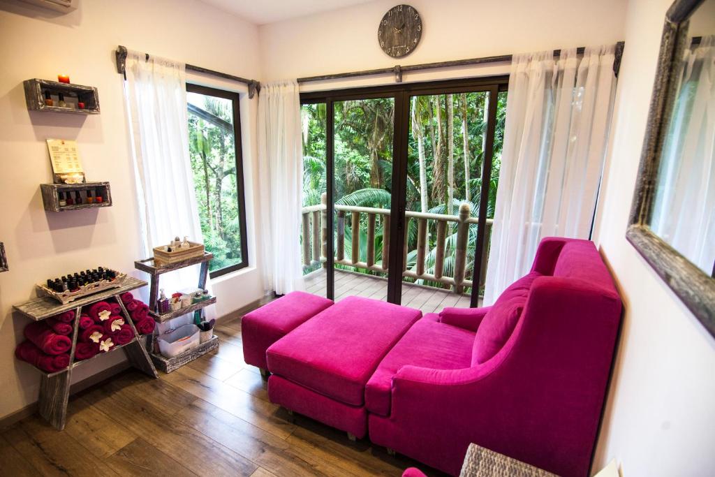 Mūsdienīgs un mājīgs spa telpa ar rozā dīvānu, koka grīdu un lieliem logiem, caur kuriem redzams zaļš meža skats. Uz plauktiem izvietotas nagu lakas un dvieļi, radot relaksējošu atmosfēru.