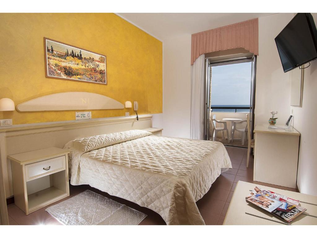 Mūsdienīga viesnīcas istaba ar dzeltenām sienām, divguļamo gultu un balkonu, kas piedāvā skatu uz jūru. Telpā ir televizors, galdiņš un maza komoda.
