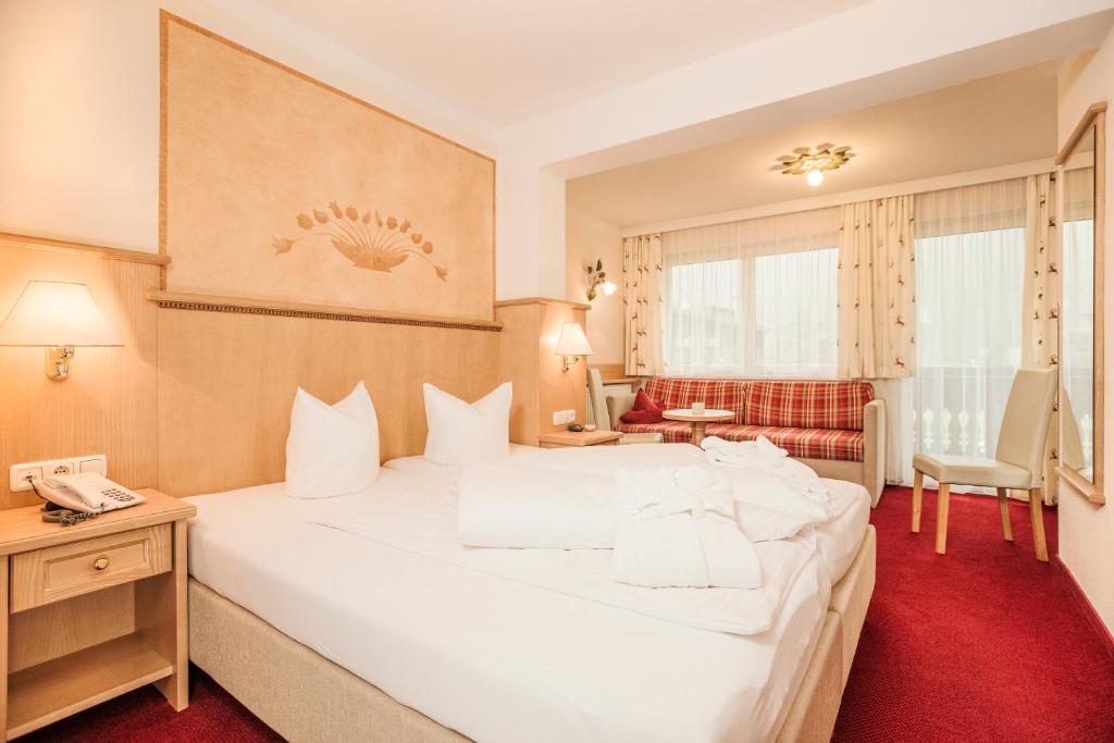 Mūsdienīga viesnīcas numura interjers ar divguļamo gultu, sarkanām paklāja grīdām un ērtu atpūtas zonu. Logi ar aizkariem nodrošina dabisko apgaismojumu.