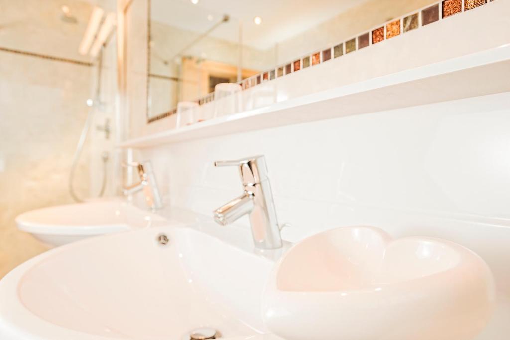 Mūsdienīga vannas istaba ar divām izlietnēm, no kurām viena ir sirds formas. Fons ir gaišs un elegants, ar modernām jaucējkrāniem un mozaīkas akcentiem.