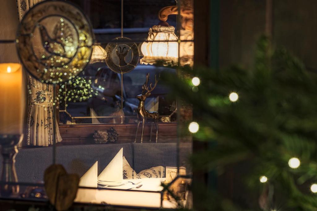 Dekoratīvs skats caur logu uz mājīgu interjeru, kur redzami sveces, Ziemassvētku rotājumi un eleganta galda uzstādīšana. Klasiskās detaļas un gaismas rada siltu un svinīgu atmosfēru.