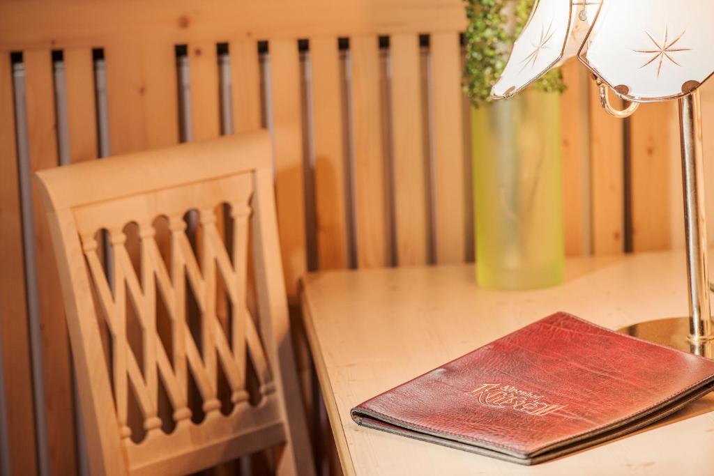 Koka galds ar sarkanu ēdienkarti un elegantu lampu, blakus atrodas koka krēsls un zaļš zieds.