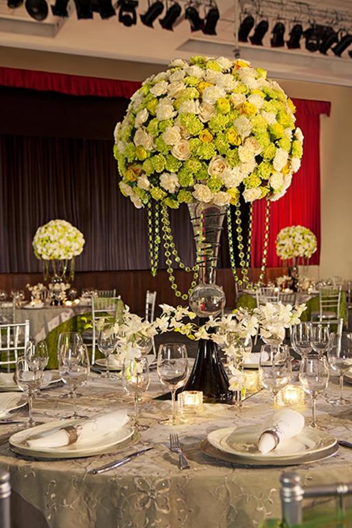 Elegants svinību galds ar grezniem ziedu centrdaļām, kas rotā pasākumu telpu. Galdu rotā sudraba trauki un glāzes, radot svinīgu atmosfēru.