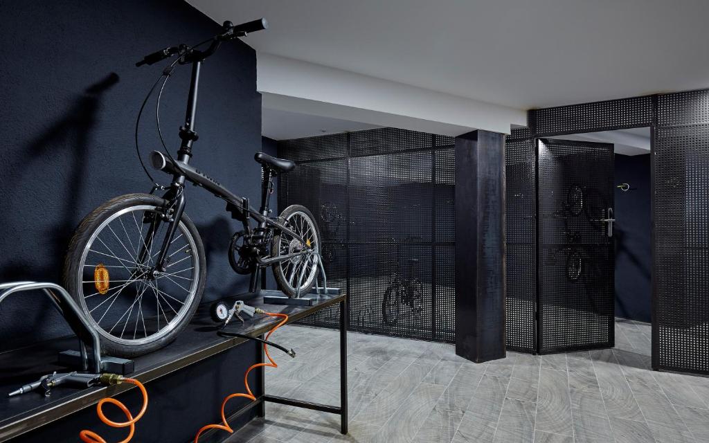 Mūsdienīga velosipēdu uzglabāšanas telpa ar melnu sienu un metāla režģa durvīm, kur ir redzami divi velosipēdi un dažādi rīki.