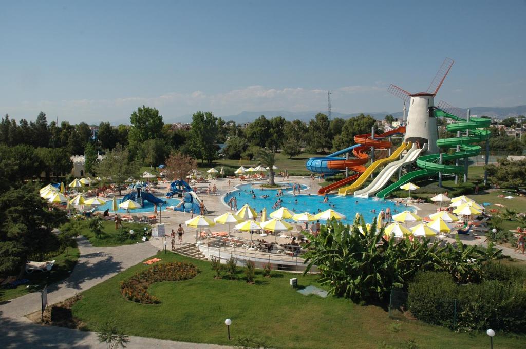 Ūdens atrakciju parks ar dažādiem slidkalniņiem un peldbaseinu, kurā ir daudz apmeklētāju. Apkārt esošie koki un sauļošanās krēsli ar dzeltenām parasolēm rada relaksējošu atmosfēru.