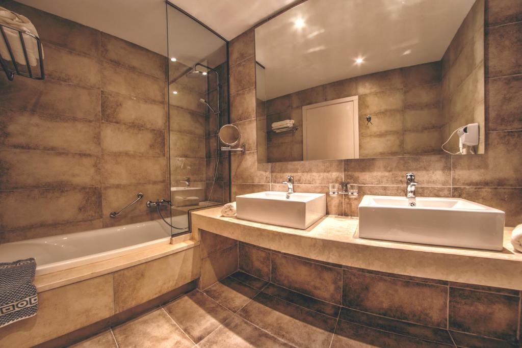 Mūsdienīgs vannas istabas interjers ar divām izlietnēm, vanna un stikla duša. Siltas krāsas akmens apdare rada relaksējošu atmosfēru.