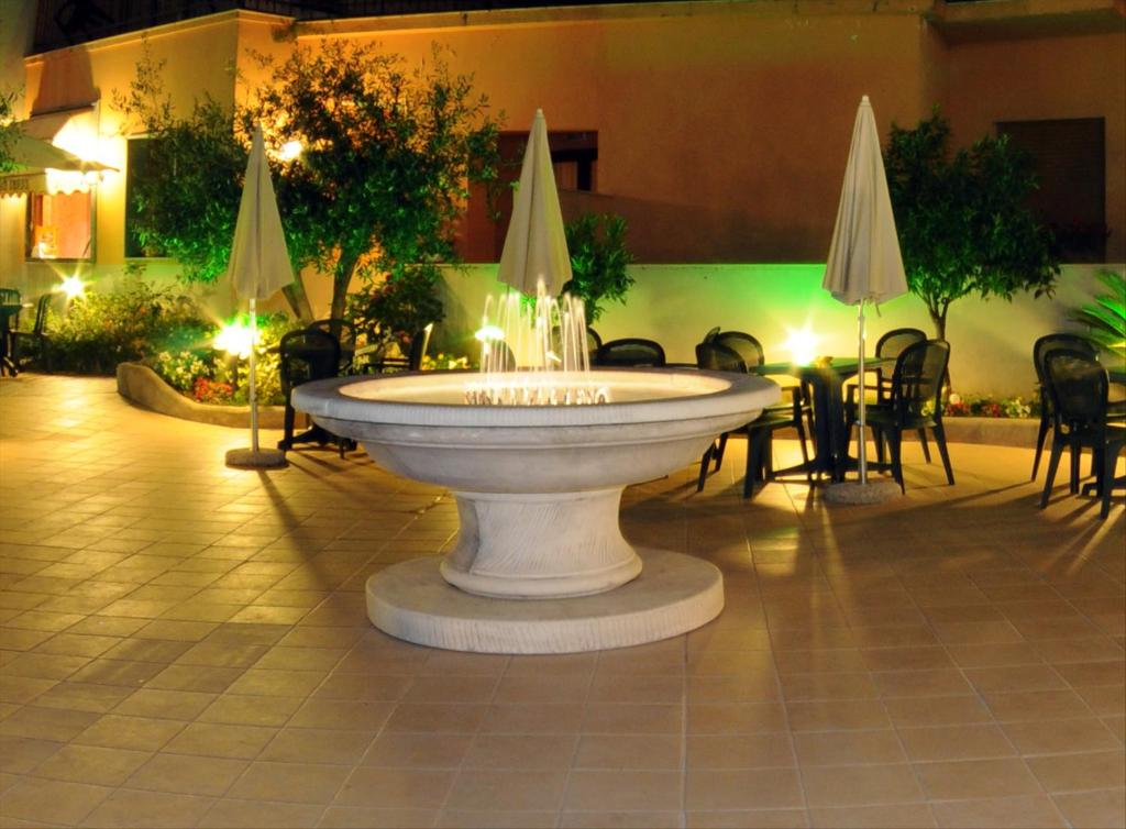 Dārza strūklaka nakts apgaismojumā, apņemta ar zaļumiem un ērtām sēdvietām. Šī vieta ir ideāla relaksācijai un vakara pavadīšanai.