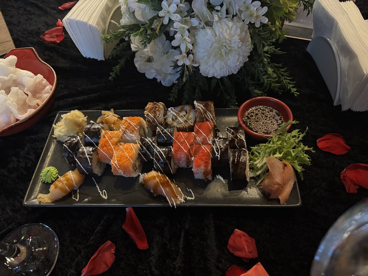 Svaigu sushi šķīvja attēls ar dažādiem rullīšiem, garnelēm un pievienotiem garnējumiem, piemēram, wasabi un ingveru. Fonā redzamas ziedu kompozīcijas un rožu ziedlapiņas. panoramic view with beach and pools
