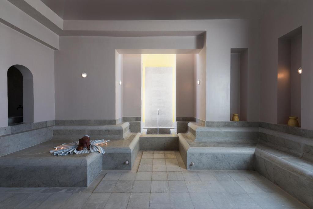 Mūsdienīga spa telpa ar akmens sēdvietām un minimālistisku dizainu, kas nodrošina mierīgu atmosfēru relaksācijai. Gaisma ieplūst caur atvērtu logu, radot patīkamu noskaņu.