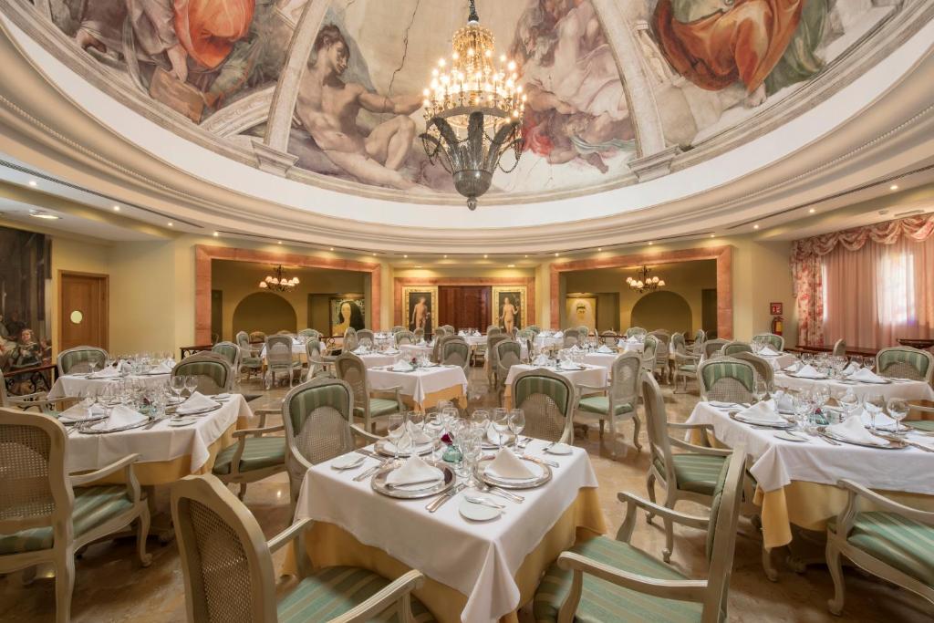 Elegants restorāns ar greznu interjeru, kurā redzami galdi ar baltiem galdautiem un elegantām trauku komplektiem. Telpas rotā skaista glezna uz griestiem un izsmalcināta apgaismojuma ķēde.