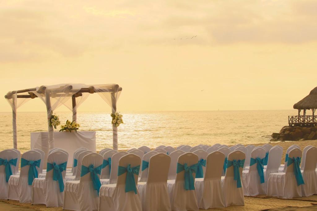 Pludmales kāzas ar baltām krēslu pārklājumiem un zila lenti, dekorēts altāris ar ziediem, saulrieta gaismā.