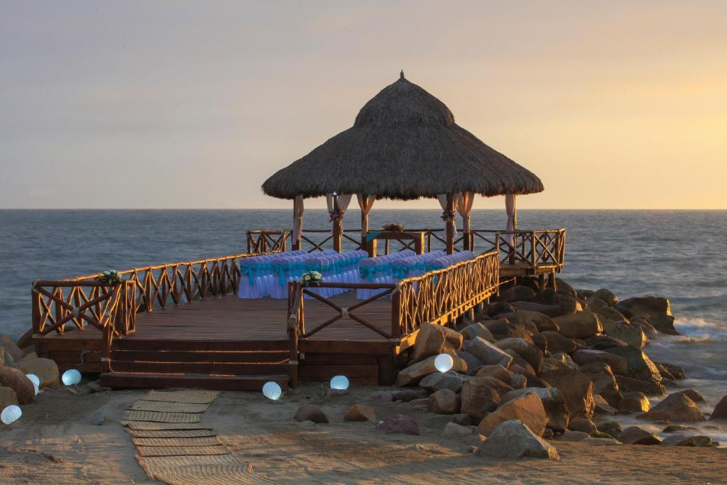 Koka piestātne ar jumtu pie jūras, kurā ir sēdvietas ar zilu apdari, radot romantisku atmosfēru saulrieta gaismā.