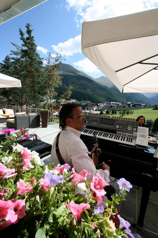 Mūziķis spēlē uz klavierēm ārpusē, ar kalnu ainavu un ziediem priekšplānā. Gaisotne ir relaksējoša, ideāla mūzikas baudīšanai.