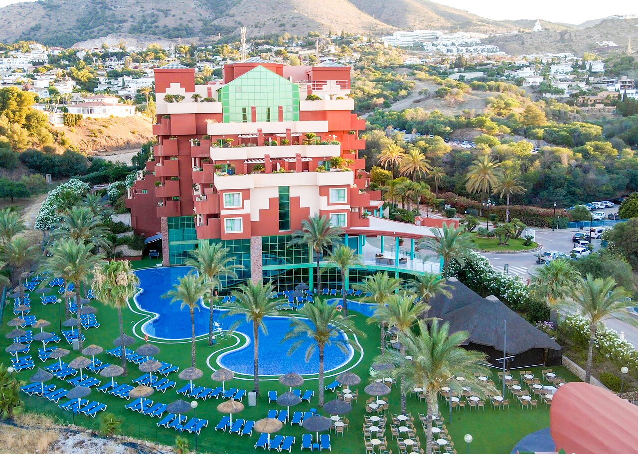 Krāsains viesnīcas komplekss ar peldbaseinu un palmām, kas atrodas kalnu ainavā. Apkārt plaša atpūtas zona ar sauļošanās krēsliem un lietussargiem. panoramic view with beach and pools