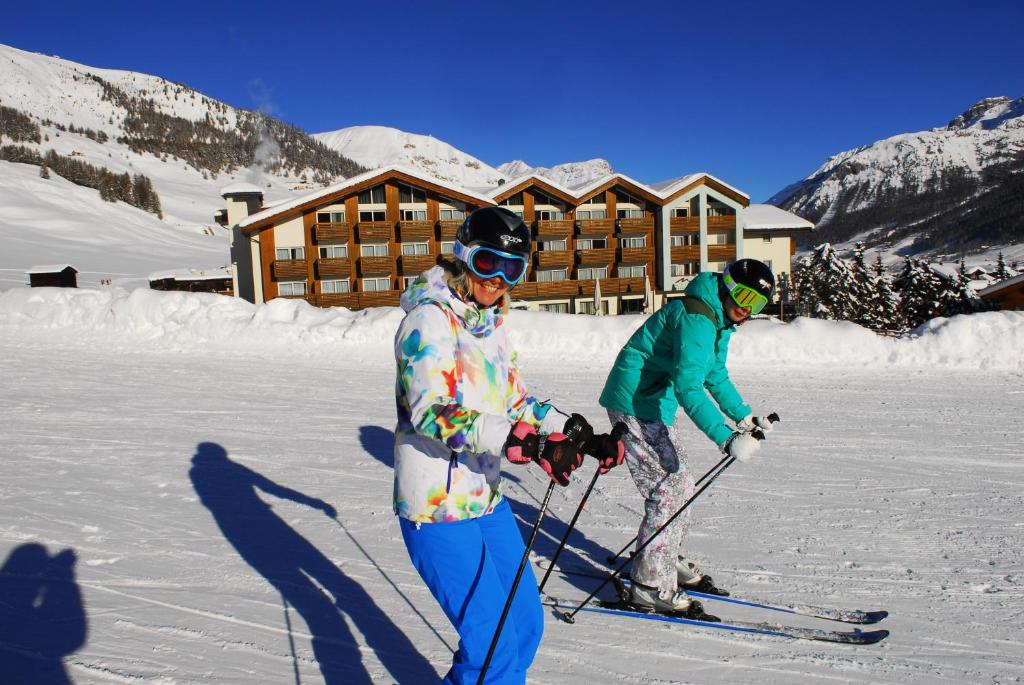 Divas slēpotājas, ģērbtas krāsainās ziemas jakās, slēpo sniegotā kalnu ainavā ar viesnīcu fona. Skaidrs laiks un kalnu virsotnes papildina ziemas sporta aktivitātes.