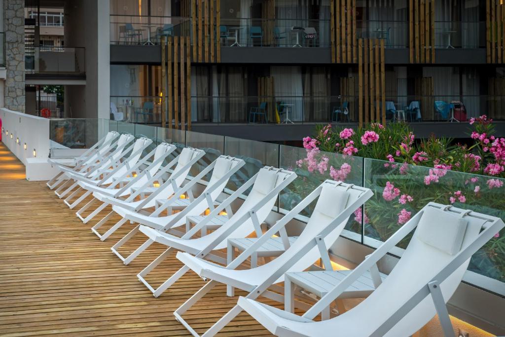 Balconā novietoti balti sauļošanās krēsli, kuriem fonā redzami ziedi un moderna arhitektūra. Apspīdēti ar gaismu, tie rada relaksējošu atmosfēru.