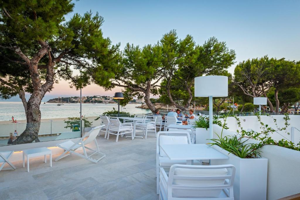 Pludmales restorāns ar modernu terasi, kurā redzami baltie galdi un krēsli, zaļie koki un skats uz jūru. Ideāla vieta relaksācijai un ēdiena baudīšanai pie ūdens.