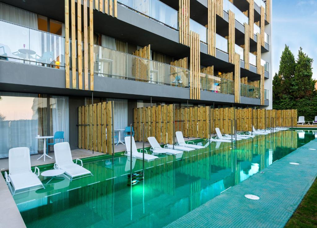 Moderns viesnīcas komplekss ar stikla balkoniem un baseinu, kurā ir sauļošanās krēsli un koka žogi. Peldbaseins atrodas pie ēkas, kas apzaļumota ar augiem.