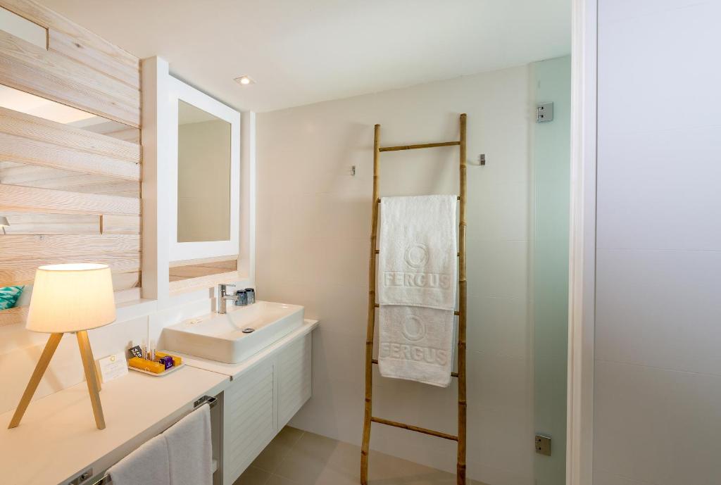 Mūsdienīgs vannas istabas interjers ar baltu izlietni, koka akcentiem un gaismas lampu. Redzama arī dvieļu turētāja un spogulis.