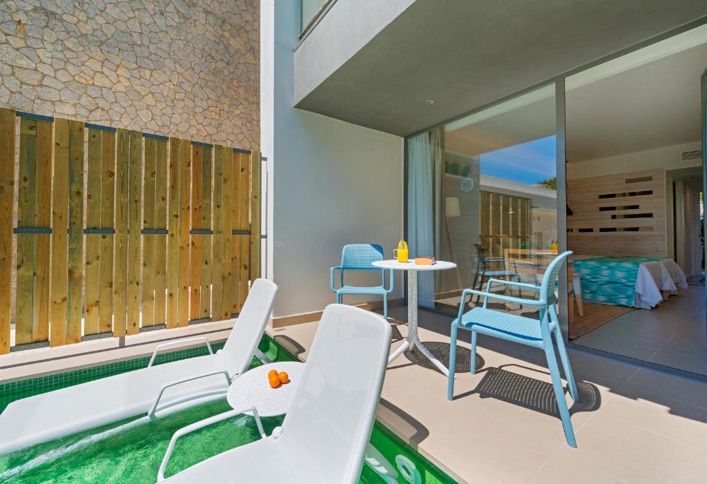 Mūsdienīga terase ar baseinu, kurā ir baltas sauļošanās krēsli un maza galdiņa komplekts, ideāli piemērota atpūtai. Fons veidots no akmens un koka, radot relaksējošu atmosfēru.