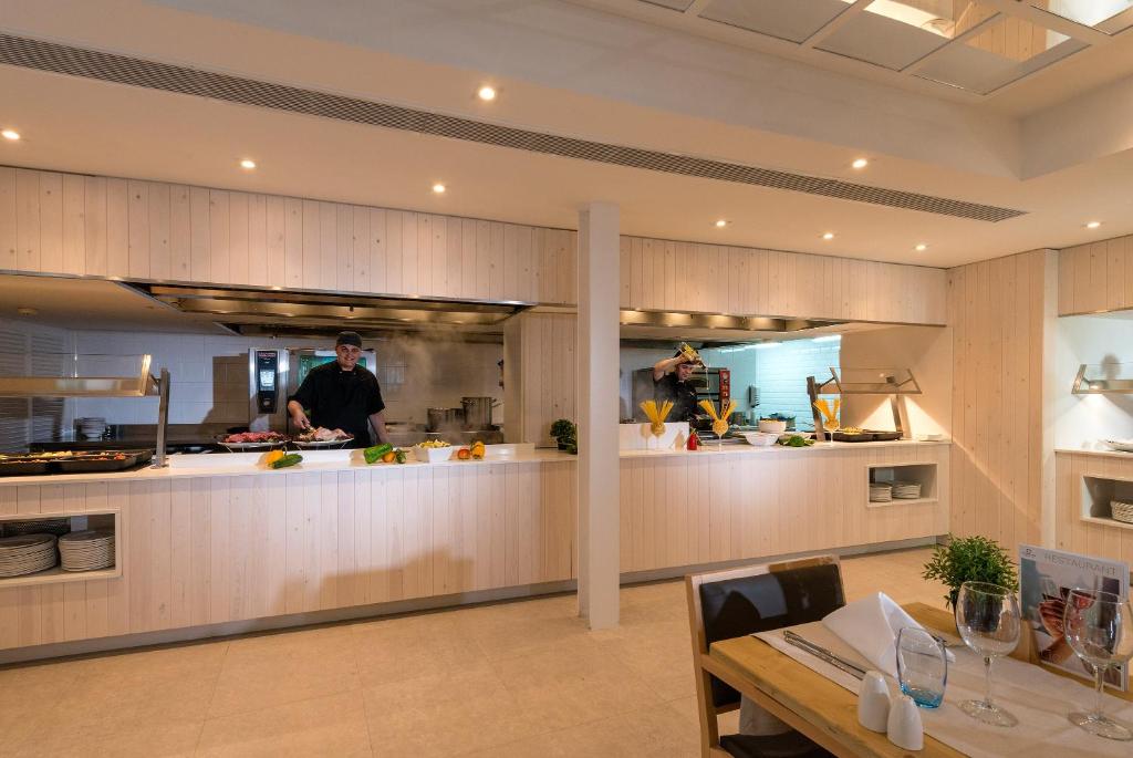 Mūsdienīga virtuve ar šefpavāriem, kas gatavo ēdienus, apgaismojuma elementiem un koka apdari. Svaigi dārzeņi un gatavošanas rīki uz galda.