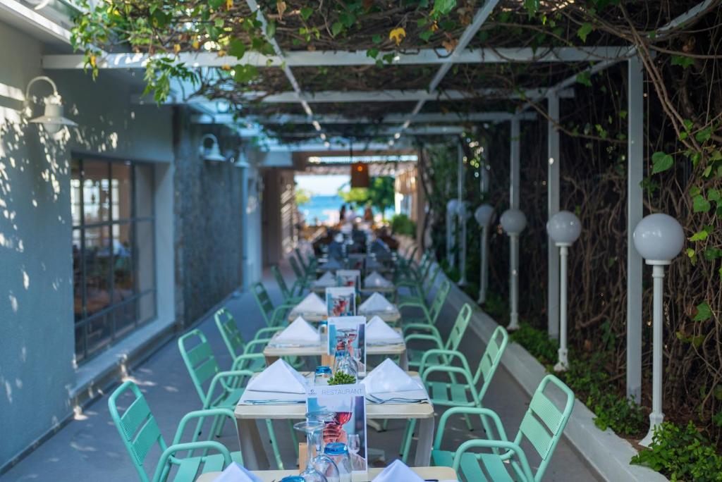 Gaismas piepildīta terase ar zaļām krēsliem un galdiem, kas klāti ar baltiem salvetēm, sniedzot patīkamu atmosfēru ēdienreizei. Dārzā redzami augi un apgaismojums, kas rada relaksējošu vidi.